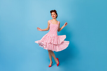 Carefree pinup girl dancing and laughing. Cute young woman in polka-dot dress having fun on blue...