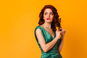 Pensive girl touching curly hair on yellow background. Studio shot of thoughtful woman in green dress.