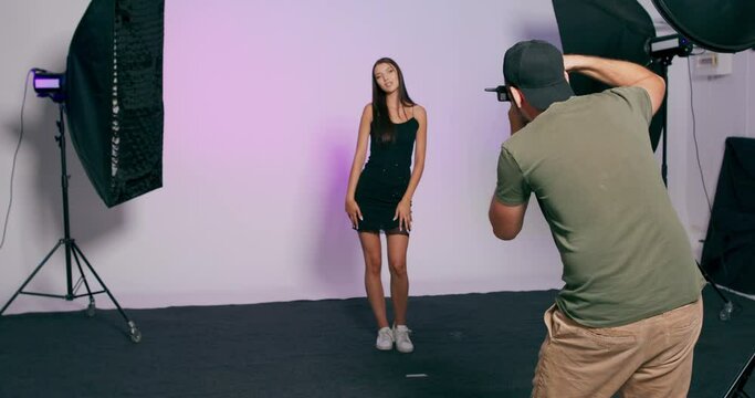 A male photographer with a baseball cap takes photos of a model against a backdrop in a studio.