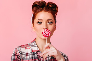 Amazed ginger girl in checkered shirt looking at camera. Studio shot of pinup woman with candy isolated on pink background.