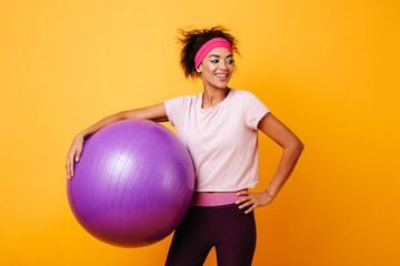 Inspired african girl posing after training on yellow background. Happy african woman holding fitness ball.