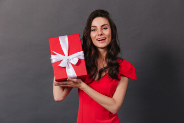 Close-up portrait of smiling dark-haired woman holding decorated box with gift. Lady in red is smiling sincerely on black background