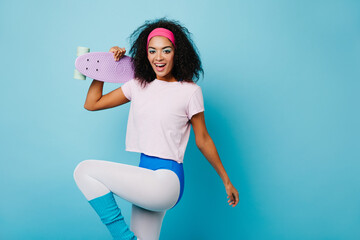 Cheerful black girl in sport attire dancing on blue background. Merry african woman with purple skateboard laughing at camera.