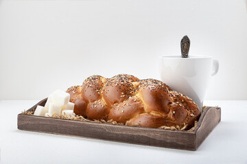 Wicker wheat bread sprinkled with sesame seeds and cereals on a wooden tray with a white mug with tea, a spoon, and lump sugar on a light background