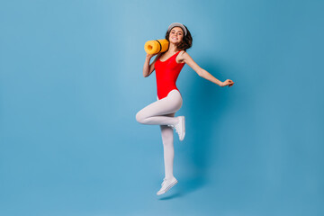 Fototapeta na wymiar Girl in tight spots suit happily jumps on blue background. 80s woman carries her yellow fitness mat