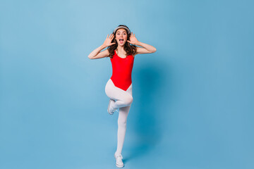 Full-length portrait of active girl in sports bodysuit and leggings. Woman jumps and demonstrates fitness exercises on blue background