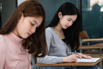 Young female university students concentrate on doing examinations in the classroom. Girl students seriously write the exercise of the examinations in the classroom.