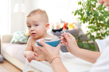 Mother's hand feeds funny cute happy blond baby toddler with spoon
