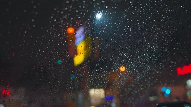 Raindrops run down the glass of the car against the backdrop of the bright lights of the night city. Rainy night. Cars pass the intersection. Bright colored traffic lights.
