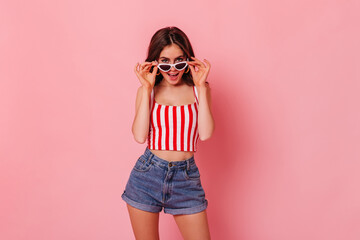 Slim girl in denim shorts and striped top puts on stylish sunglasses on pink background