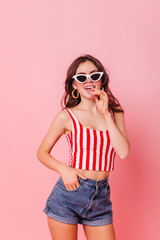 Fototapeta premium Young girl in blue shorts and red-white top coquettishly posing on pink background. Brunette with glasses and gold earrings looks into camera