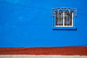 A window covered with an ornate iron cover in a blue wall with brick texture at the bottom.