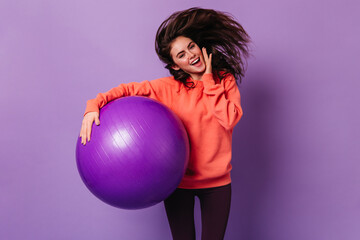 Smiling lady in bright sweatshirt and dark leggings jumps on purple background, holding fitball