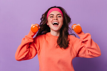 Perky girl in sports outfit holding dumbbells. Curly dark-haired lady engaged in fitness on purple...
