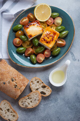 Turquoise plate with baked feta cheese, tomatoes, green olives and ciabatta bread, top view on a light-blue stone surface, vertical shot