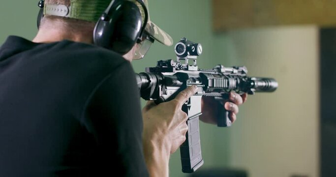 Back view of a man wearing ear protection shooting a rifle in an indoor firing range.