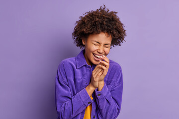 Overjoyed curly haired woman keeps hands together laughs out from positive emotions dressed in...