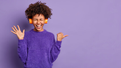 Overjoyed curly haired woman raises palm feels very happy points thumb away on copy space area wears casual jumper stereo headphones enjoys music isolated over purple background. Look at this
