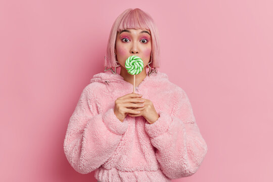 Glamour gorgeous Asian lady with bright makeup covers mouth with round green lollipop wears pink hair wig and fur coat poses indoor. Mllennial girl holds caramel candy on stick has sweet tooth