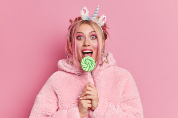 Pretty European woman has makeup licks delicious candy looks surprisingly at camera wears...