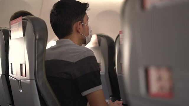Brunet young man with protective mask sits on chair in light cabin of contemporary comfortable airplane close side view slow motion.