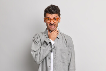 People negative emotions health problems concept. Displeased man keeps hand on throat suffers suffocation clenches teeth from unpleasant feelings wears spectacles and grey shirt poses indoor