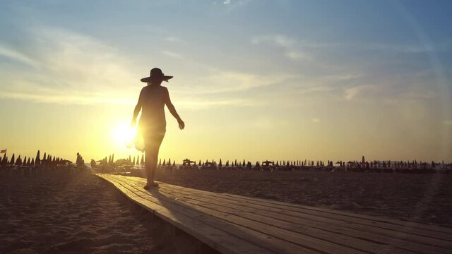 Rear view of young beautiful romantic brunette woman with dress and hat walking on wooden boardwalk walkway on beach toward sunset. Peace of mind, freedom and travel concept