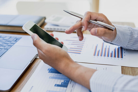 A Businessman Is Using A Mobile Phone To Compare Information With The Documents On His Desk. He Is Working In His Office.