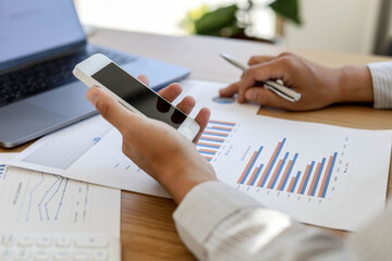 A businessman is using a mobile phone to compare information with the documents on his desk. He is...