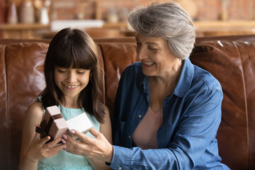 Close up smiling mature older grandmother presenting gift to cute little granddaughter, sitting on couch at home, happy curious girl opening box, family celebrating birthday, two generations
