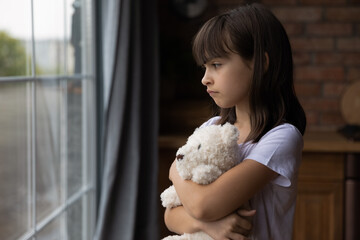 Close up lonely little girl hugging toy, looking out window, standing at home alone, upset unhappy...