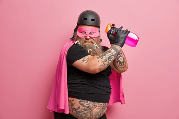 Funny bearded man with big tattooed belly wears helmet eyemask cloak holds cleaning detergent going...