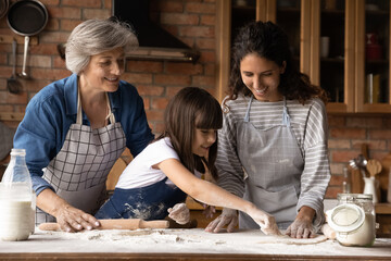 Happy three generations of women cooking handmade pastry, kneading, rolling dough, standing in kitchen, happy little girl with smiling mother and mature grandmother enjoying leisure time at home