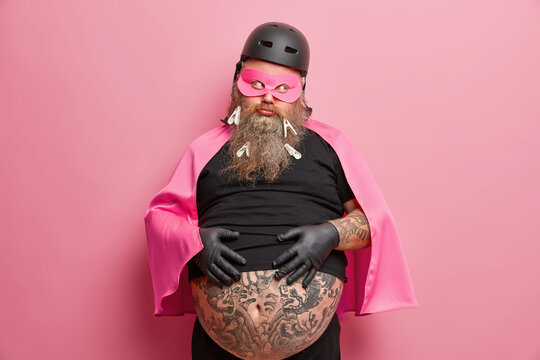Sad unhappy bearded European man keeps hands on tattooed belly has discontent face expression bored look dressed in superhero costume isolated over pink background. Overweight male hero indoor