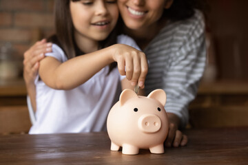 Close up smiling mother and little daughter hugging, putting coin into pink piggy bank, caring mum...