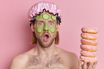 Headshot of surprised man with bristle keeps mouth opened applies green cucumber mask wears water...