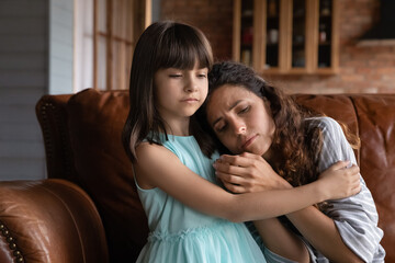 Close up caring little girl comforting, hugging depressed mother, sitting on couch at home, preschool daughter calming, embracing upset frustrated mum, family overcoming problems together, divorce