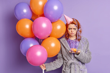 Fototapeta na wymiar Unhappy angry redhead birthday girl smeared with serpentine spray has leaked makeup holds cupcake bad mood during celebration dressed in domestic clothes holds colorful balloons. Holiday concept