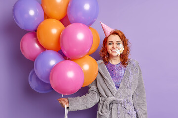 Fototapeta na wymiar Pleasant looking cheerful birthday girl gets cake on face accepts unexpected surprise wears party cone hat and gown holds inflated balloons poses against purple background. Festive occasion concept