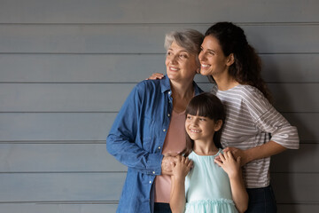 Close up three generations of women dreaming, looking to aside, standing on grey wooden wall...
