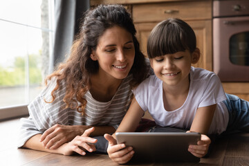 Close up happy mother and adorable little daughter playing tablet, lying on warm wooden floor with underfloor heating, enjoying leisure time with gadget, watching video, chatting or shopping online