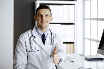 Friendly male doctor sitting and showing OK sign at his working place in clinic. Perfect medical service in hospital. Medicine and healthcare concept