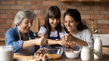 Happy little girl with smiling young mother and mature grandmother preparing dough close up, stirring fresh eggs with whisk, sitting at table in modern kitchen, family enjoying leisure time together