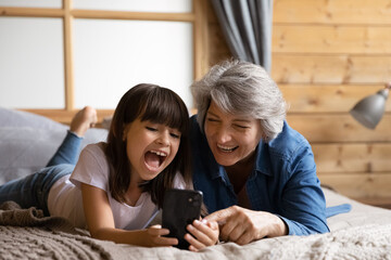 Close up overjoyed little girl with mature grandmother using phone together, lying on cozy bed at home, spending leisure time with gadget, having fun, browsing apps, chatting online, watching video