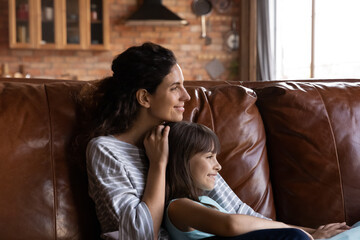 Close up dreamy mother with little daughter hugging, looking to aside, dreaming, sitting on couch at home, smiling mum and adorable girl child relaxing, visualizing future, enjoying weekend