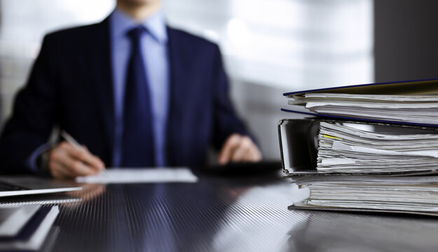Binders with papers are waiting to be processed by man accountant in blue blazer staying at home during covid pandemic. Taxes and audit concept. Internal Revenue Service inspector checking financial