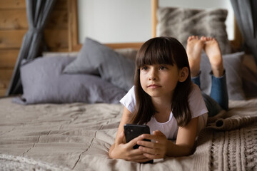 Close up dreamy little girl holding phone, relaxing, lying on cozy bed at home, adorable preschool...