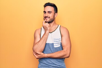 Young handsome man with beard wearing sleeveless t-shirt standing over yellow background smiling looking confident at the camera with crossed arms and hand on chin. Thinking positive.