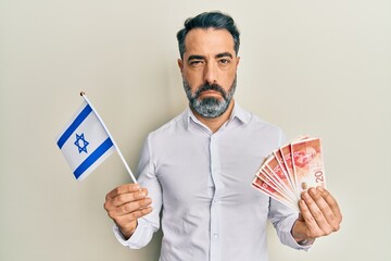 Middle age man with beard and grey hair holding israel flag and shekels banknotes depressed and...
