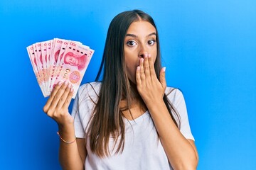 Young hispanic woman holding yuan chinese banknotes covering mouth with hand, shocked and afraid...